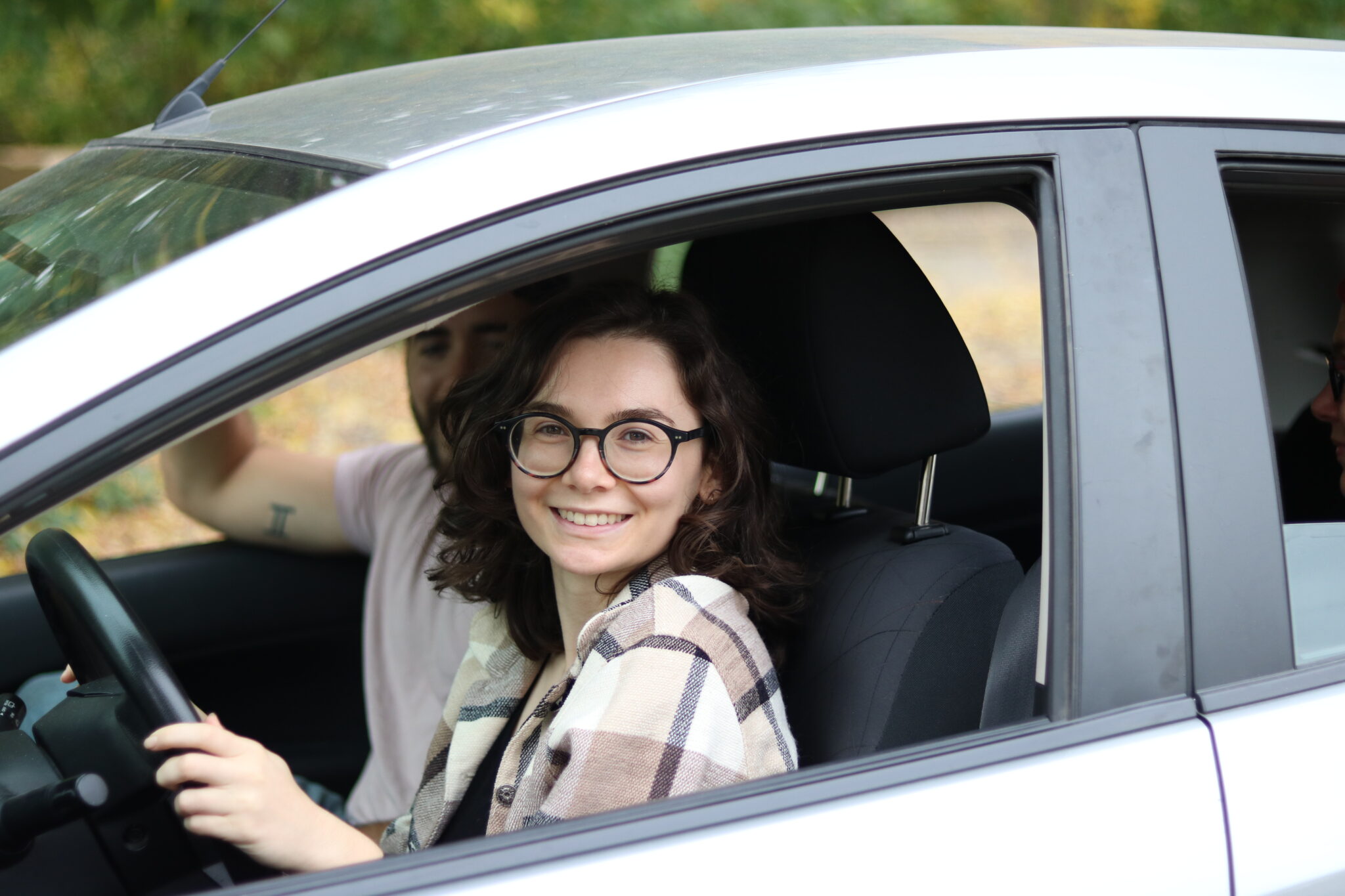 We are looking into the driver's window of a grey car. Inside is a young woman driver who is smiling at the camera.