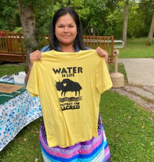 A young Indigenous woman posing with her yellow 'Water is Life' t-shirt, with a screen printed bison design. 