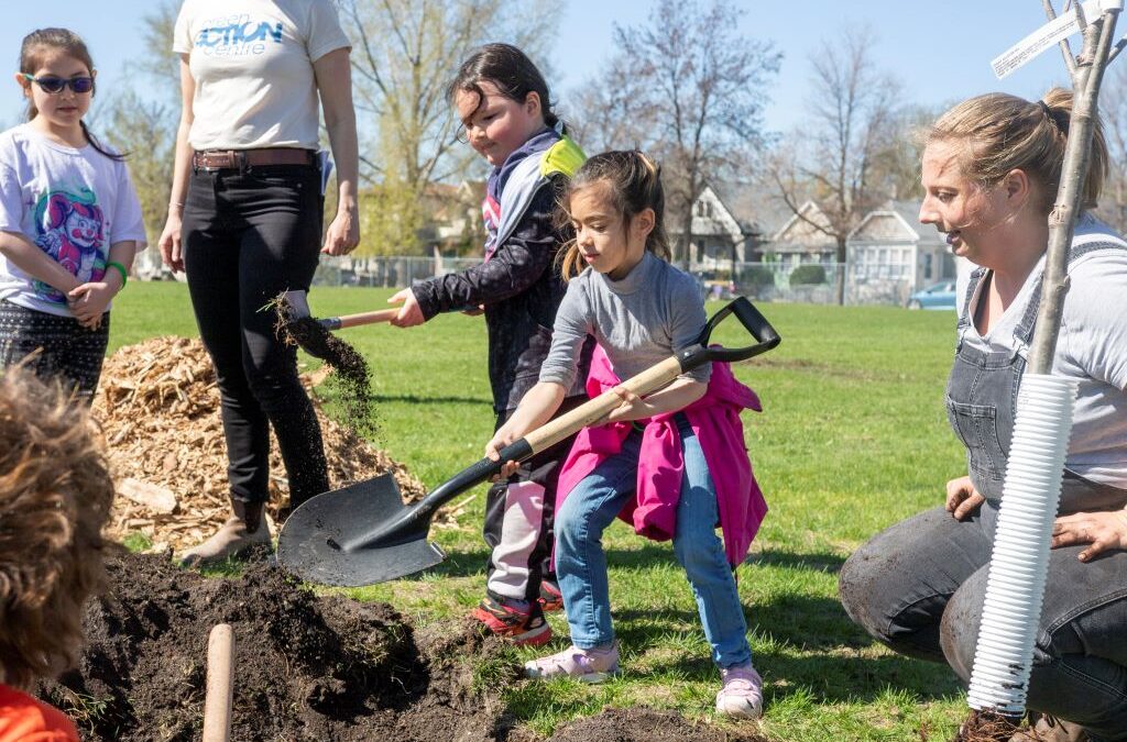 Student holding a shovel about to take a scoop from a pile of dirt. Beside the student is an adult sitting next to a small tree that's ready to be planted. There are two more students in the background, and an adult wearing a Green Action Centre t-shirt.