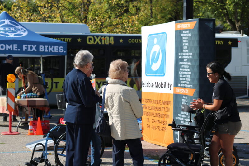 Two older adults listen to a female presenter with a mobility device in front of her. In the background of the scene, a large poster for Reliable Mobility and a Winnipeg Transit bus can be seen.