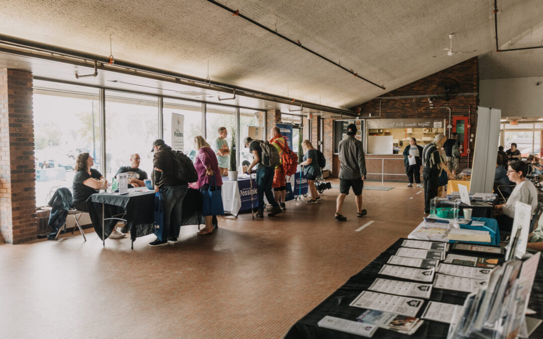 Several long tables are positioned on opposite sides of an indoor room, manned by various people in groups of one or two. Large bay windows on the opposite side of the room provide additional lighting. 5 guests speak with the vendor tables or move between tables.