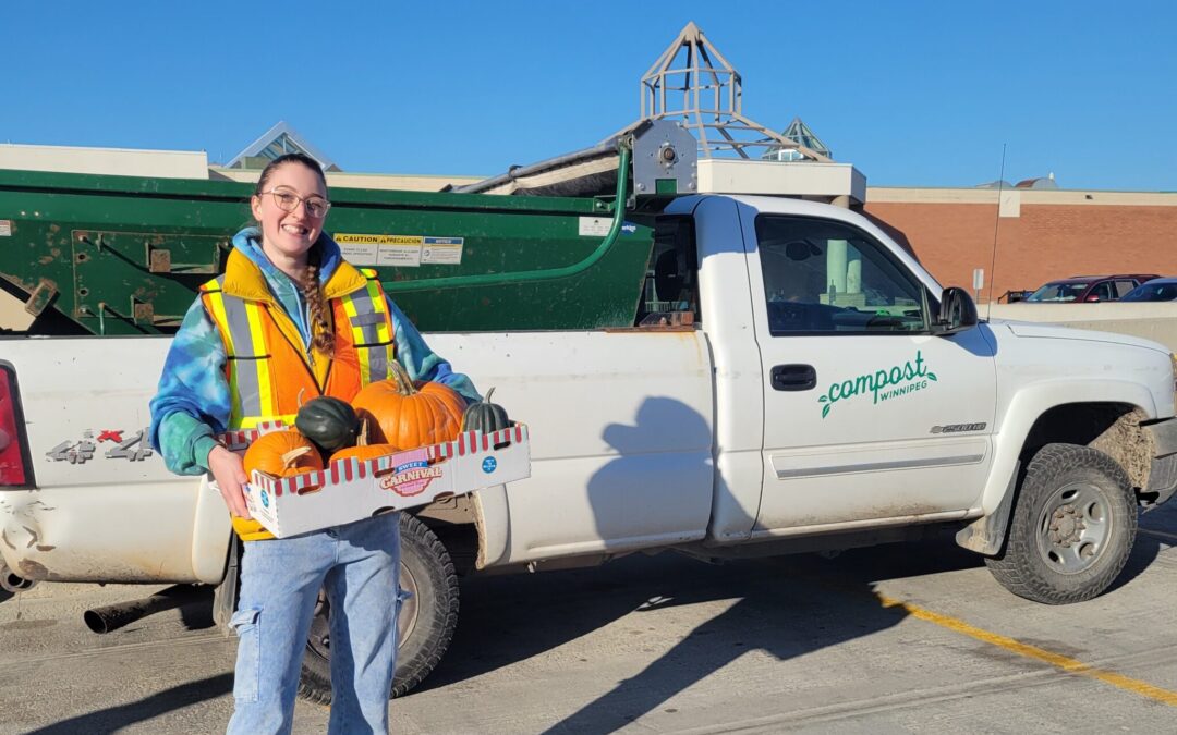 Picture of a woman in a reflective vest holding a box of pumpkins while standing in front of a white truck with the Compost Winnipeg logo on it. Behind the truck is a blue sky and the roof of the Polo Park building.