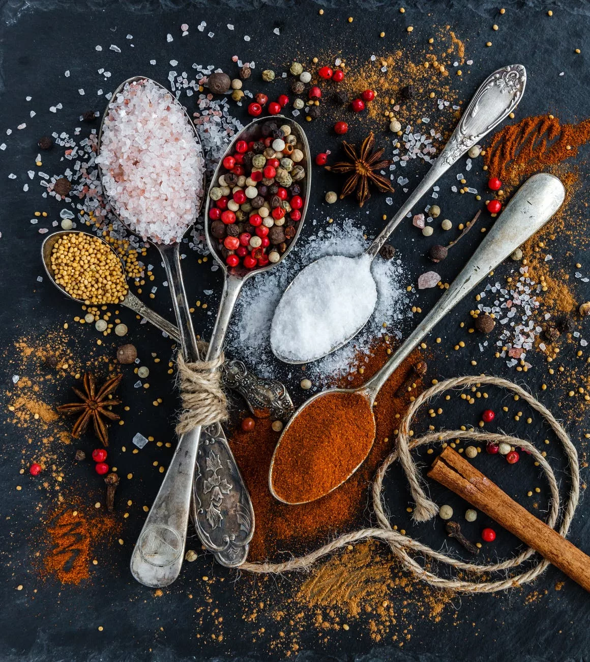 various seasonings, cinnamon, sugar, cloves, star anise, pink salt, etc on spoons and scattered across the surface