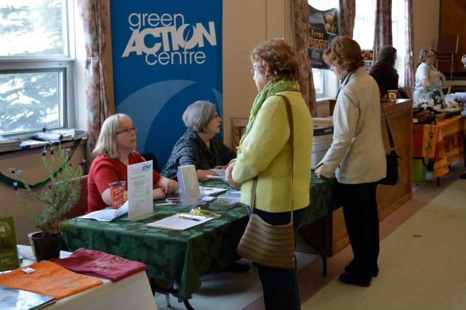 Two volunteers sitting at a table with the Green Action Centre banner in the background. They are in a conference room with other tables and vendors speaking to event attendees.