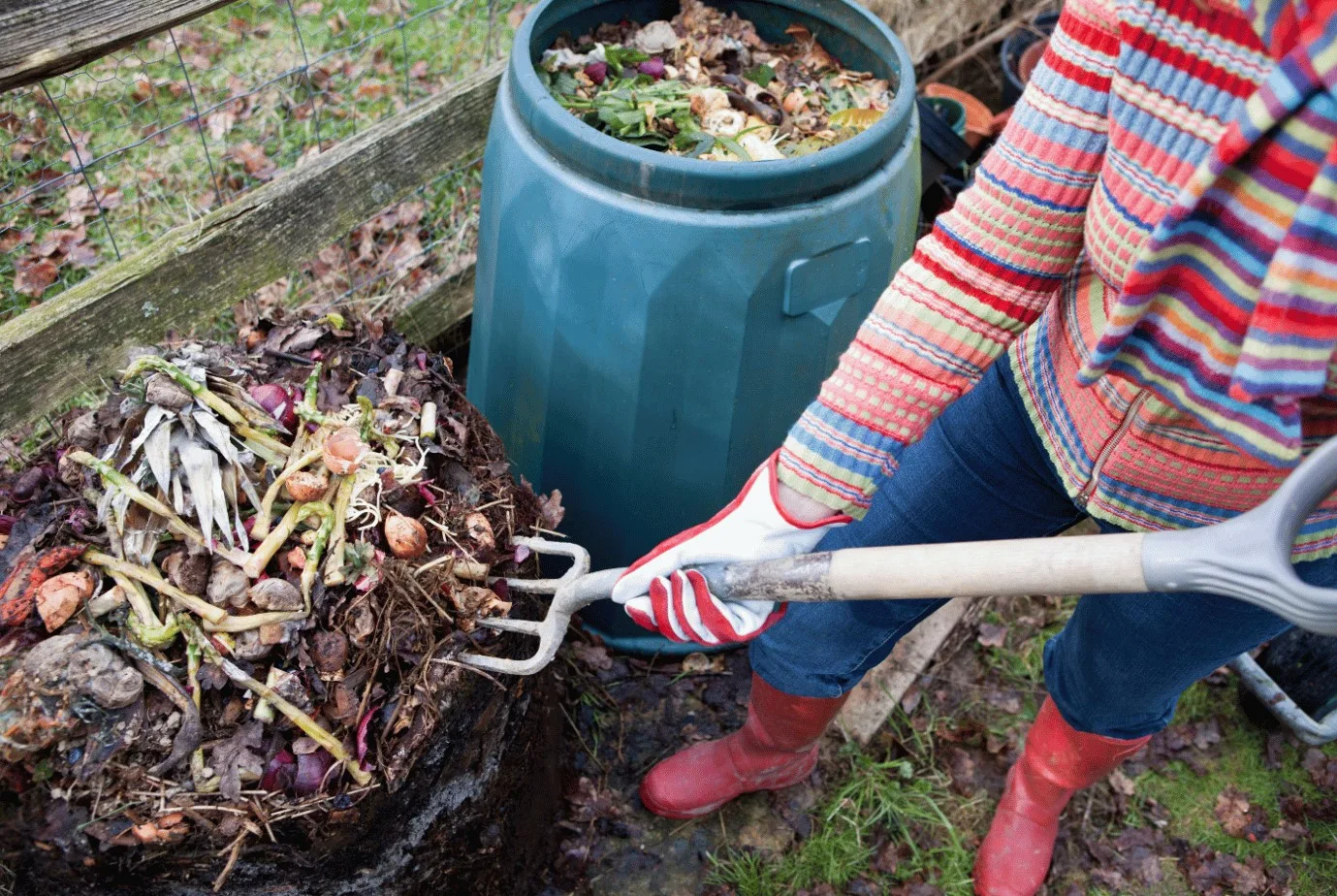 Person holding a pitchfork shoveling a compost pile beside a plastic compost bin.